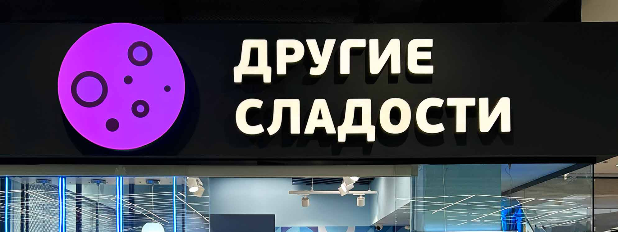 Световая вывеска «Другие сладости» с фиолетовым логотипом над входом
